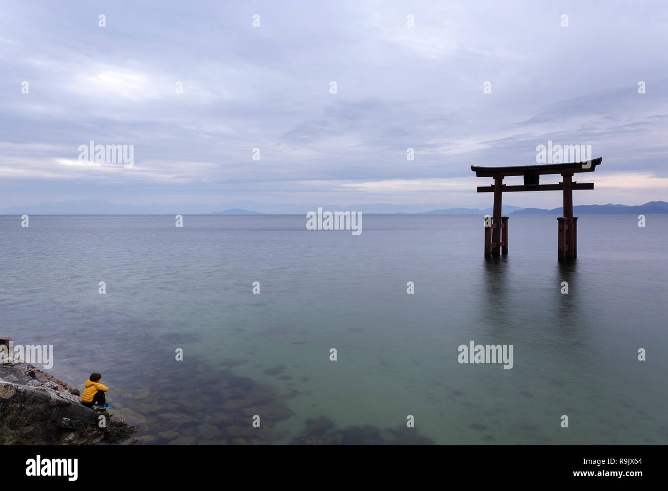 Young boy wearing yellow jacket sitting on steps on the lake looking at Torii gate on water, Shirahige Jinja Torii, lake Biwa near Kyoto, Japan Stock Photo