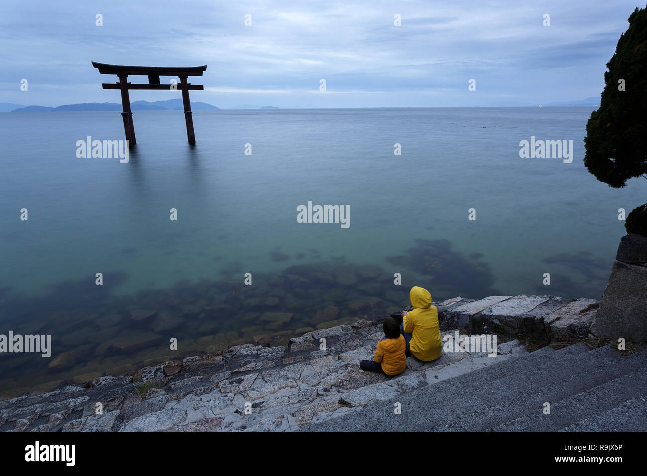 Mother and son wearing yellow jackets sitting on steps on the lake looking at Torii gate on water, Shirahige Jinja Torii, lake Biwa near Kyoto, Japan Stock Photo