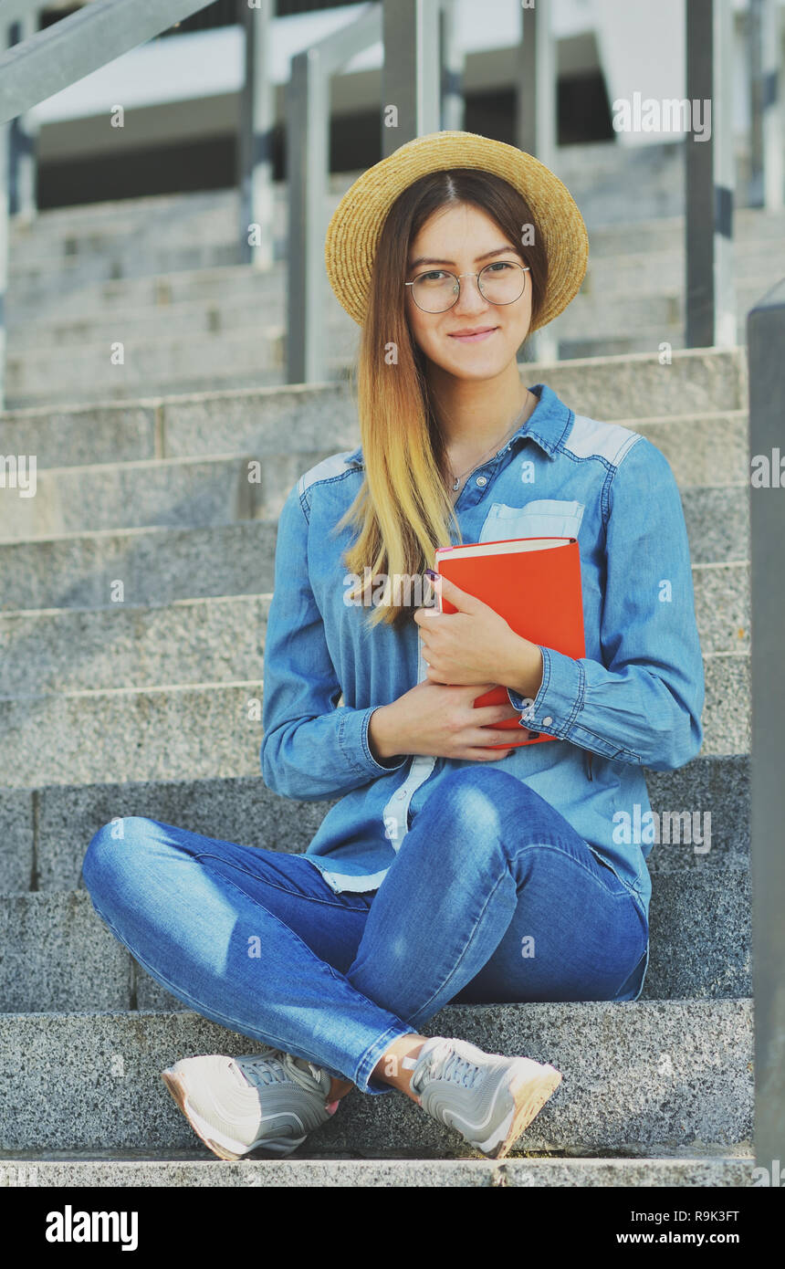 Portrait of a young girl with glasses who is wearing a straw hat and denim shirt, sitting on the steps of the campus Stock Photo