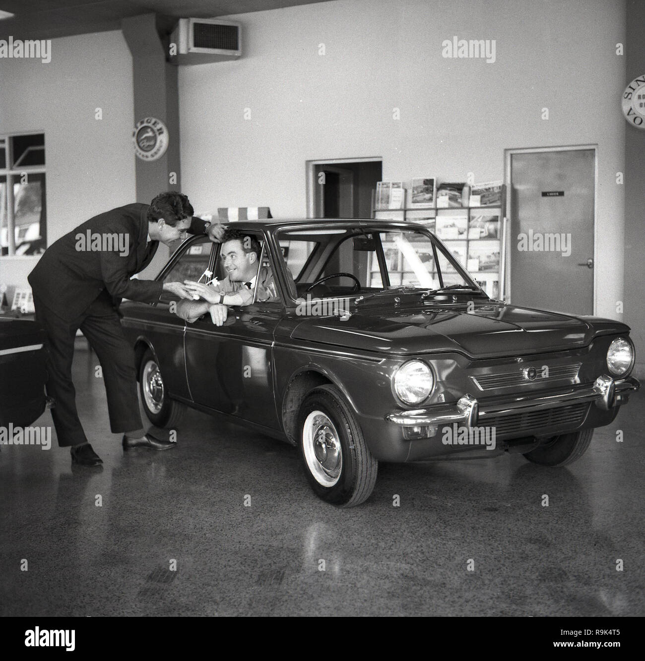 1965, historical, a man in Singer car showroom sitting in a Singer (Hillman) Imp car, England, UK being handed some large keys. The 2-door small economy car was built by the Rootes Group to compete with BMC's popular 'Mini' and featured several innovative design elements for it's day, such as a space saving aluminium rear-engine, with rear-wheel-drive. The 'Singer' Imps were badge-engineered Hillmans aimed at the slightly more upmarket small car buyer.  But reliability problems plagued the car and its losses saw the British Rootes Group taken over by the American Chrysler Corporation in 1967. Stock Photo