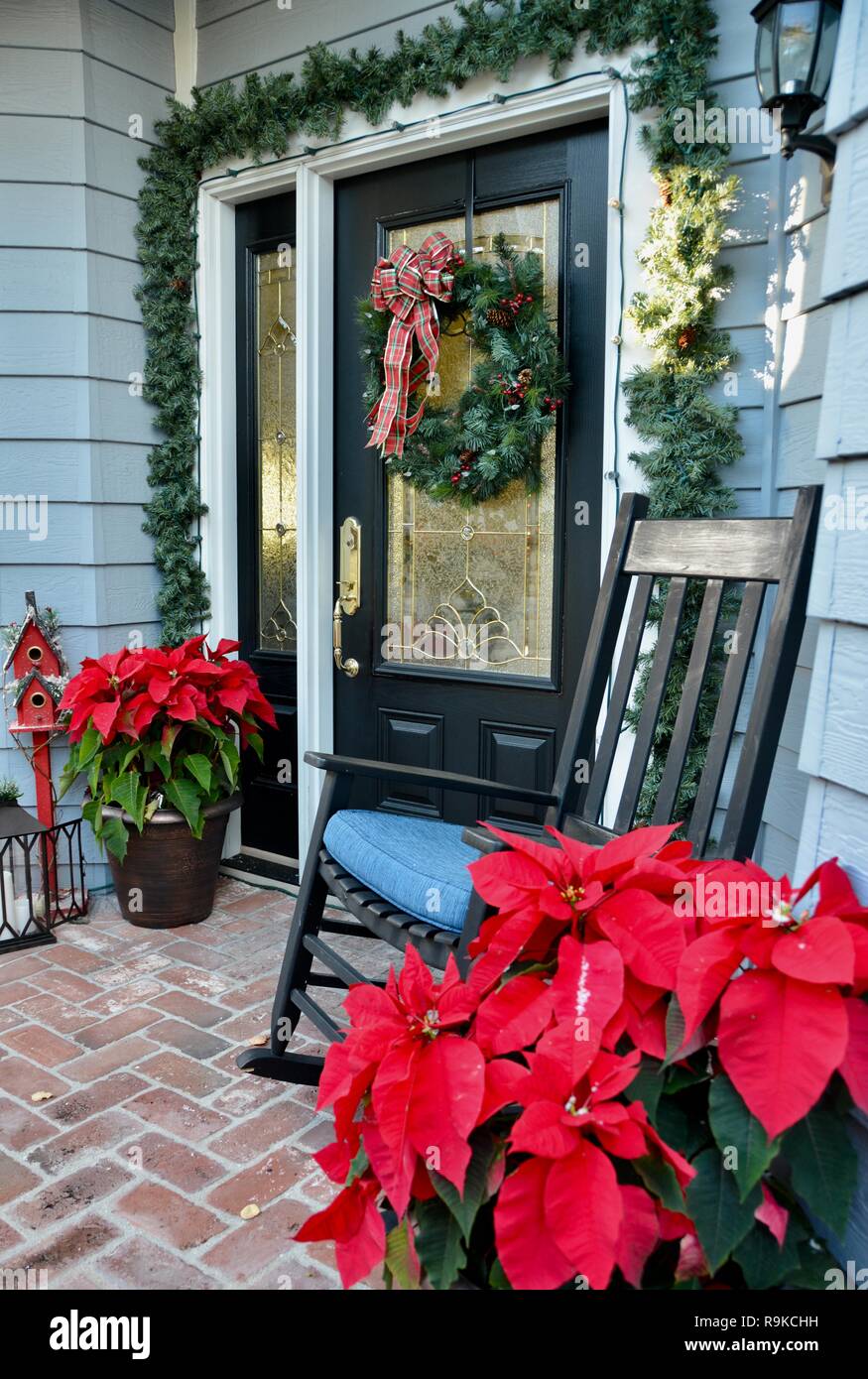 Front door entryway decorated with traditional American Christmas decor: poinsettias, wreath, lanterns, red birdhouse, evergreens and a rocking chair. Stock Photo