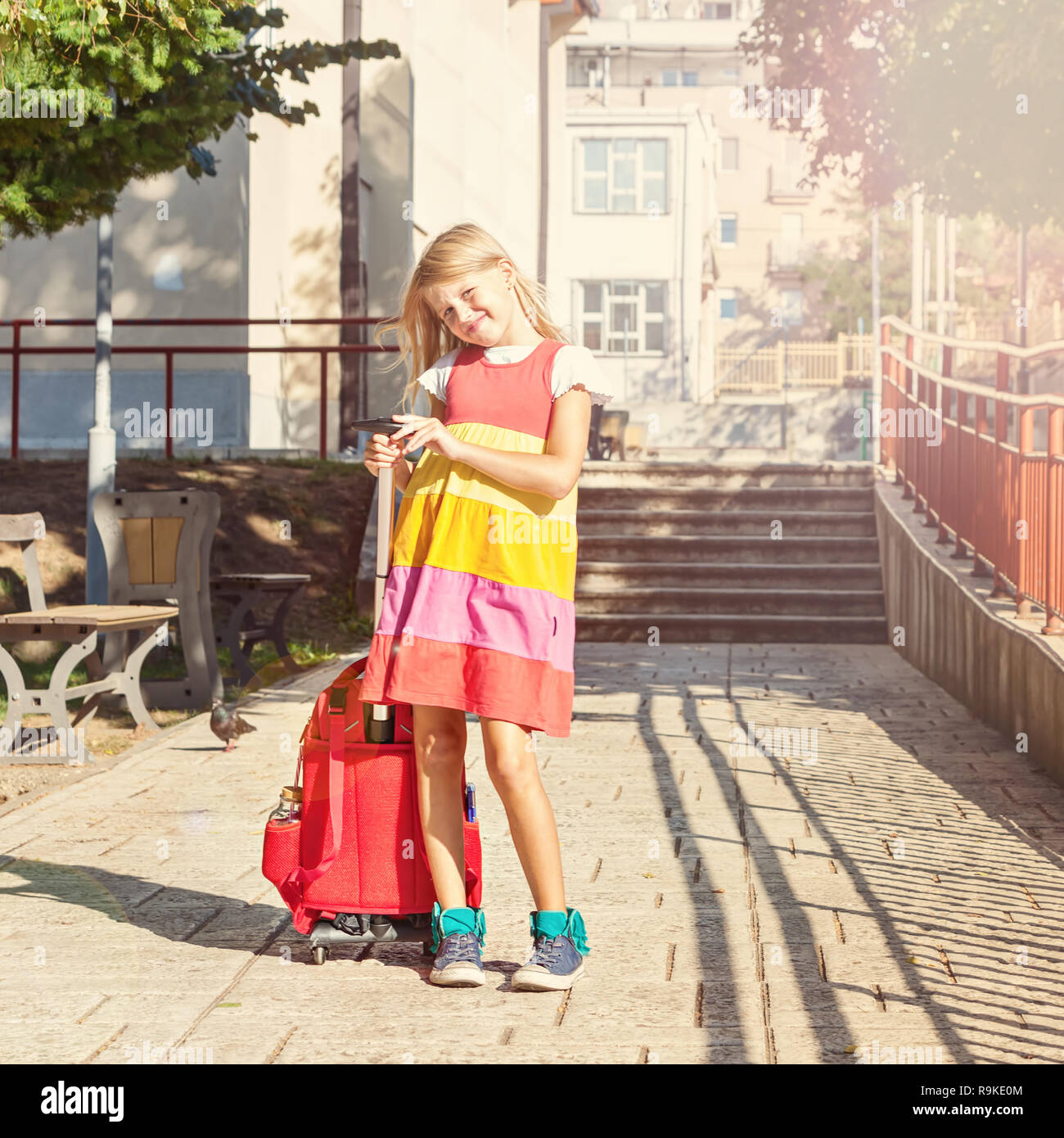 Cute 8 years old schoolgirl carries a backpack with wheels in the schoolyard Stock Photo