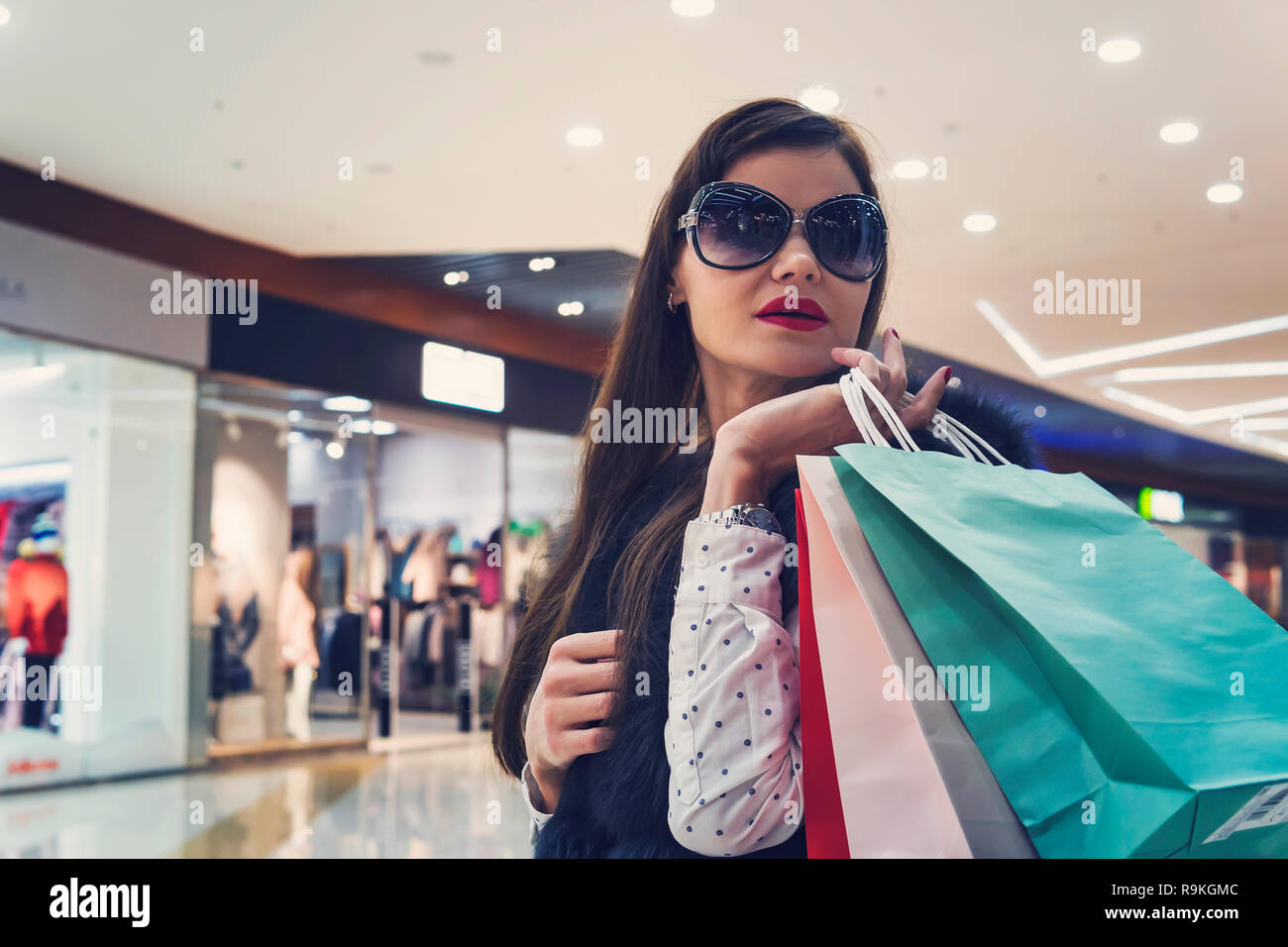 Brunette girl in dark glasses holding heap of shopping bags at mall background. Young attractive girl with red lipstick after purchasing in store. Wom Stock Photo