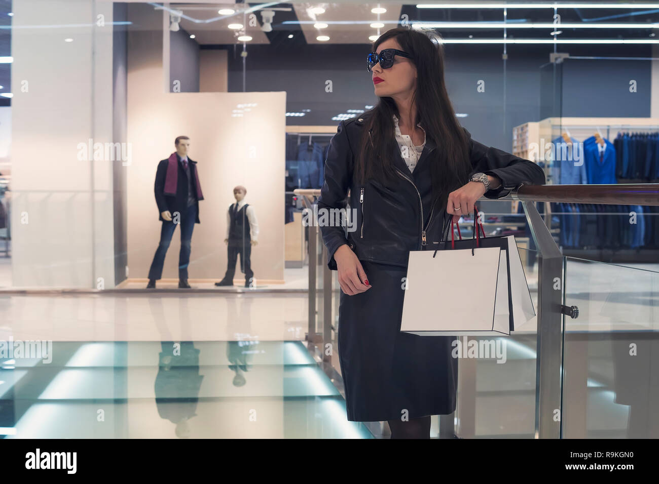 Dark-haired attractive girl standing alone on glass floor near handrail at empty shopping centre background. Lady in glasses holding white bags and lo Stock Photo