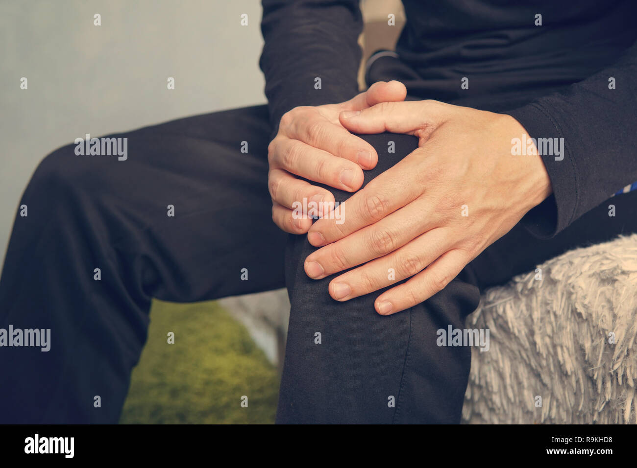 elderly man sitting on a sofa in the living room at home and touching his knee by the pain. man suffering from knee pain sitting sofa. Stock Photo