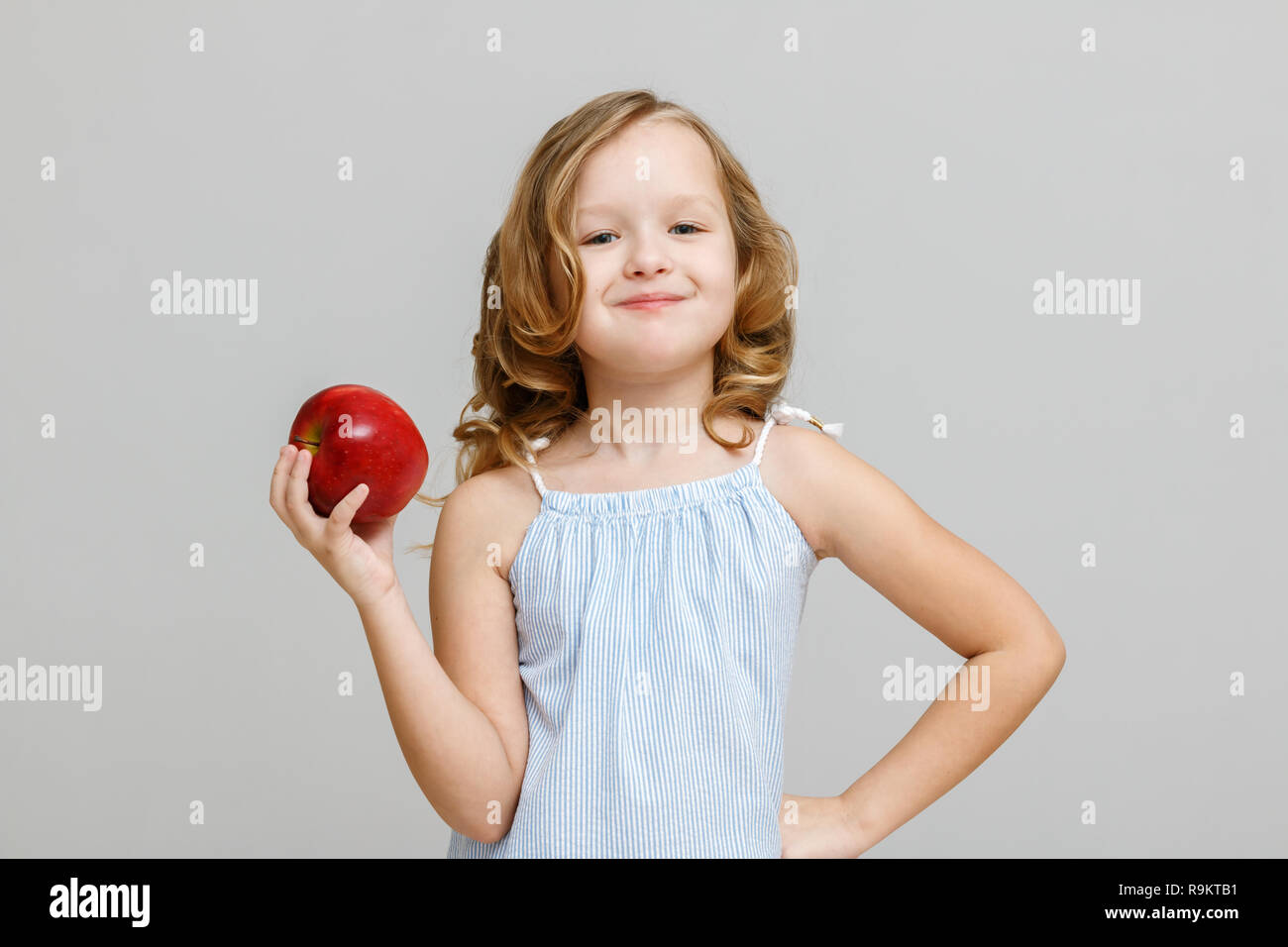 Portrait of a happy smiling little blonde girl on a gray background. Baby eating red apple Stock Photo