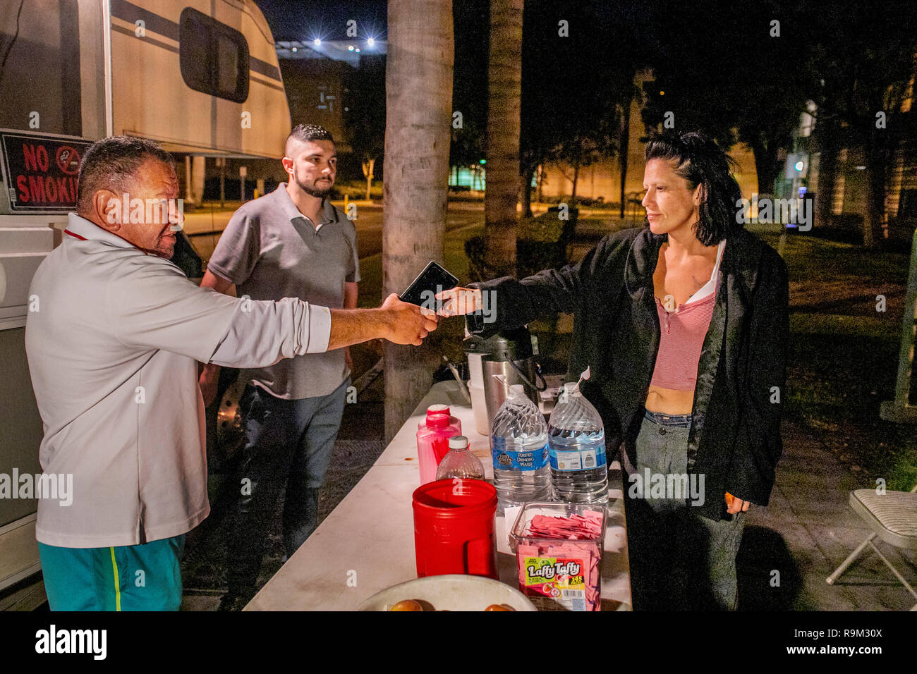 Outside a specialized van, Caucasian and Hispanic volunteers serve free food and supply cell phones to released county jail inmates on a street at night in Santa Ana, CA. Stock Photo