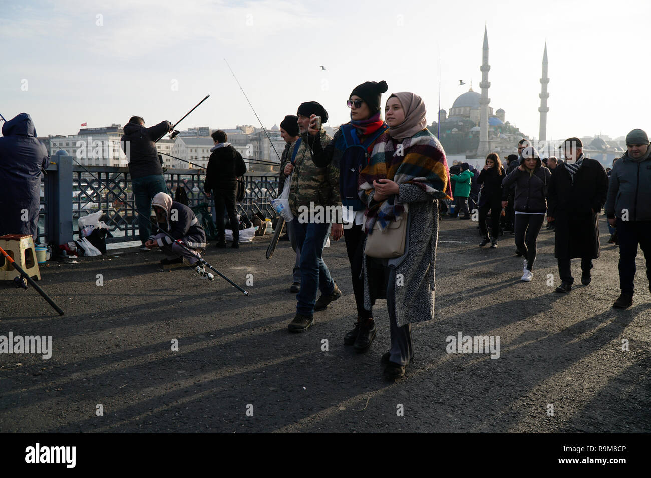 Istanbul, Turkey - December 22, 2018 : Two young Turkish Women are walking on The Galata Bridge. One of them is taking selfie.Other people are fishing Stock Photo