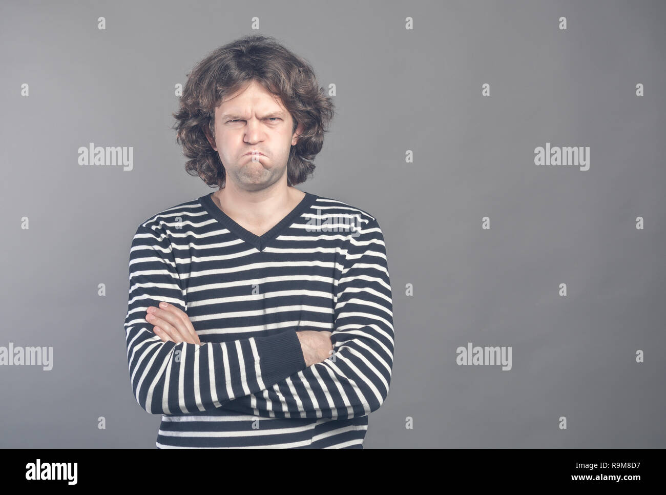 Angry young man in striped sweater puffed up his cheeks and crossed arms about to have nervous breakdown, isolated on gray wall background. Negative h Stock Photo