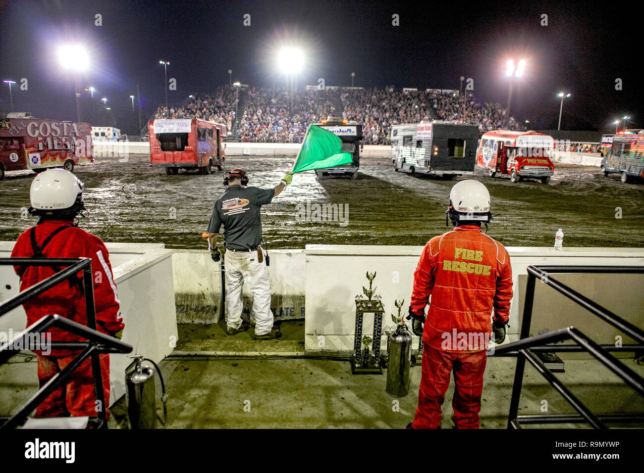 A referee waves a green flag to signal the start of a motor home-crashing demo derby in a Costa Mesa, CA, stadium. In foreground are rescue workers in safety clothing. Stock Photo
