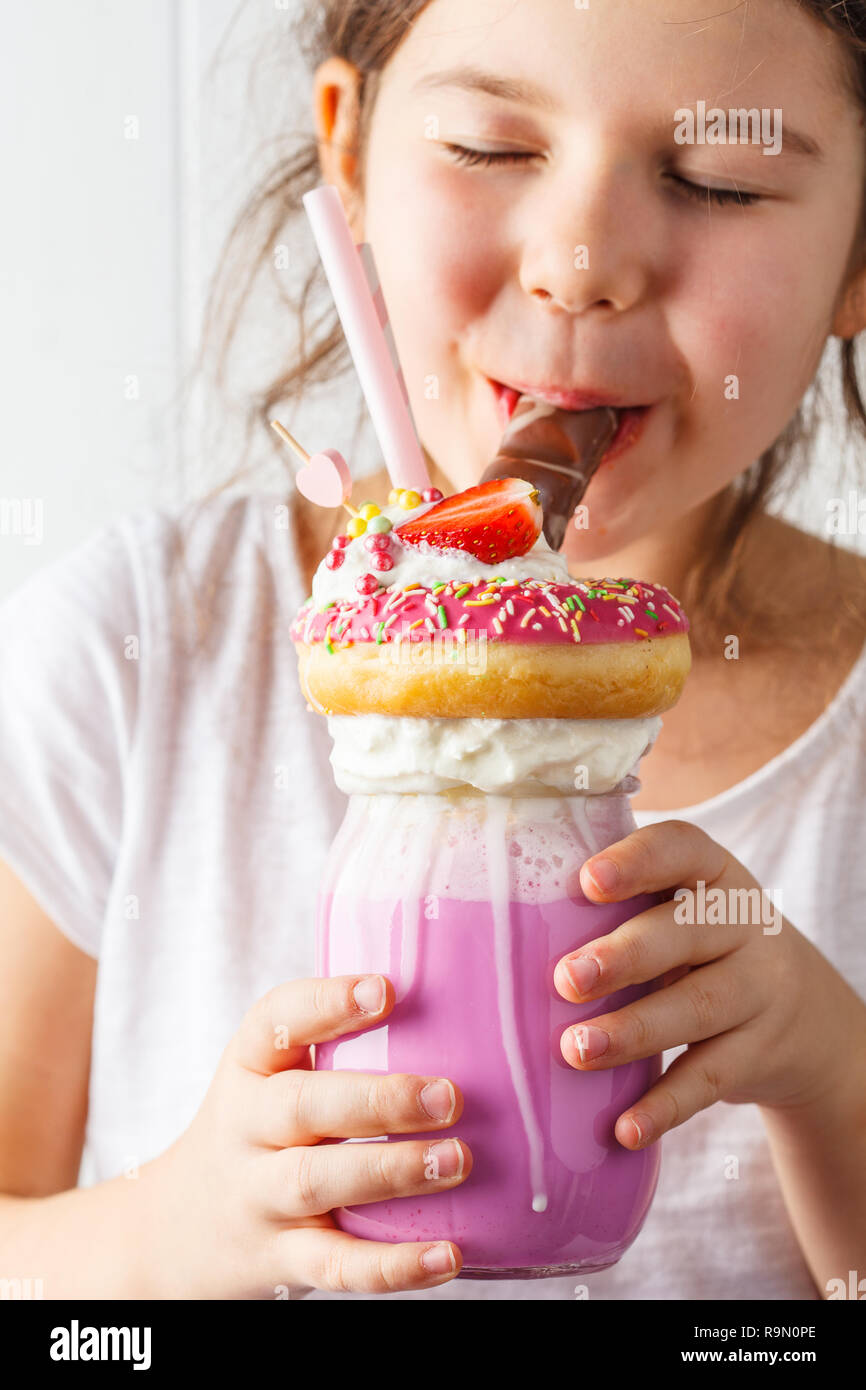 Little smiling girl drinks pink strawberry freak shake cocktail with donut and sweets, white