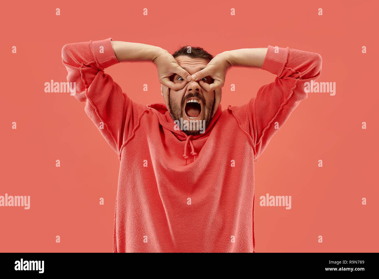 I lost my mind. The squint eyed man with weird expression. Beautiful male half-length portrait isolated on coral studio backgroud. The crazy man. The human emotions, facial expression concept. Stock Photo
