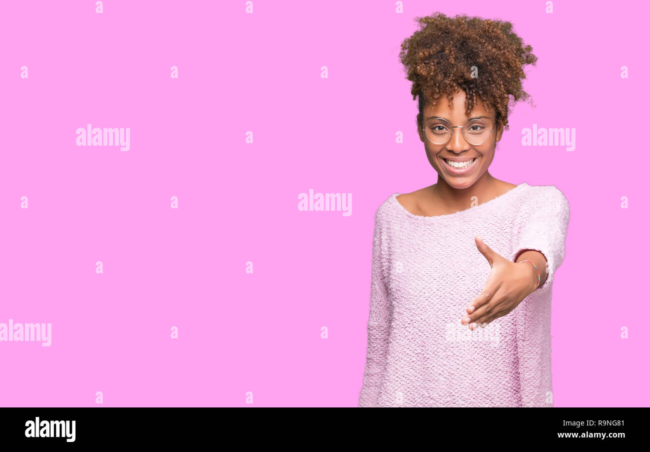 Beautiful young african american woman wearing glasses over isolated background smiling friendly offering handshake as greeting and welcoming. Success Stock Photo