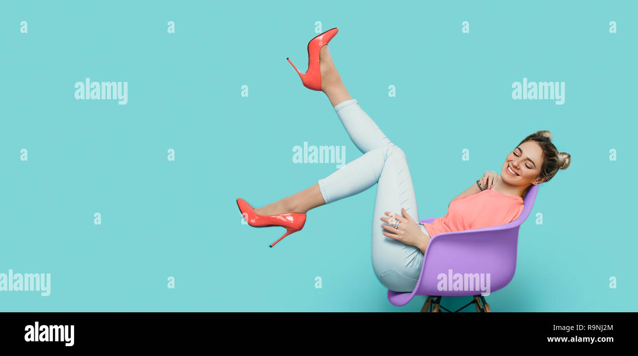 woman wearing living coral color high heel sitting on chair with raised legs against blue wall Stock Photo