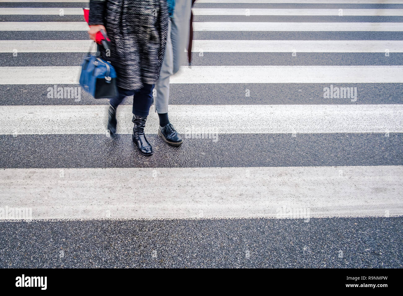 two unrecognizable persons in black shoes cross wet street after rain on crosswalk, red umbrella, parallel lines Stock Photo