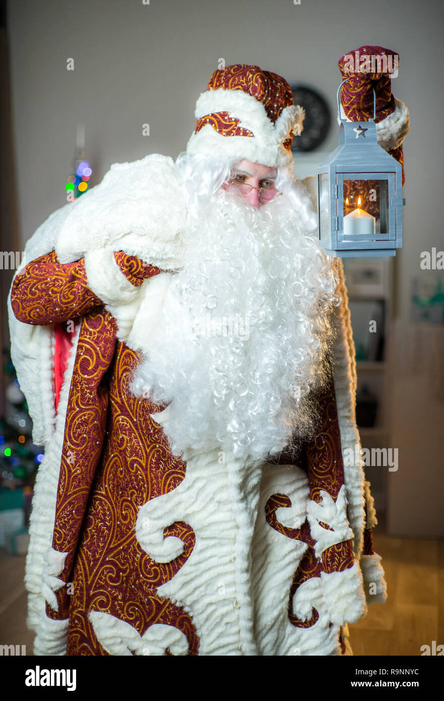 Santa Claus in a red suit. Santa with a candle lantern and gifts in his hands against the background of the New Year tree. Stock Photo