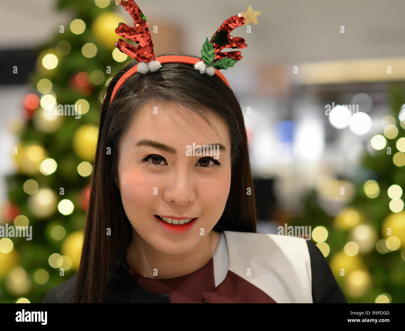 Young Thai saleslady with a Christmas headband poses for the camera in front of a decorated Christmas tree. Stock Photo