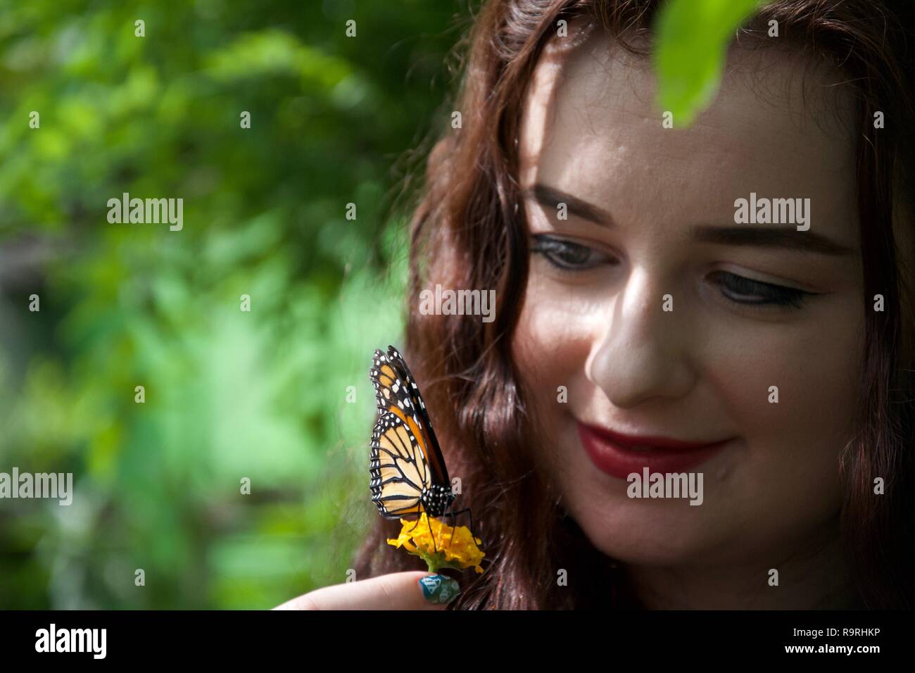A happy young lady holds a flower near her face with an orange and black butterfly sitting on it with folded wings Stock Photo