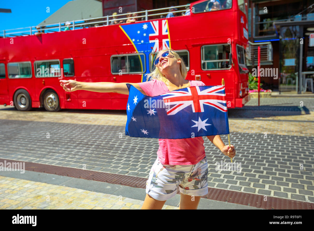 Blonde woman with Australian flag at Barrack Square near Bell Tower and Elizabeth Quay in Perth, Western Australia. Happy girl enjoys Perth downtown. Iconic open tourist bus on blurred background. Stock Photo