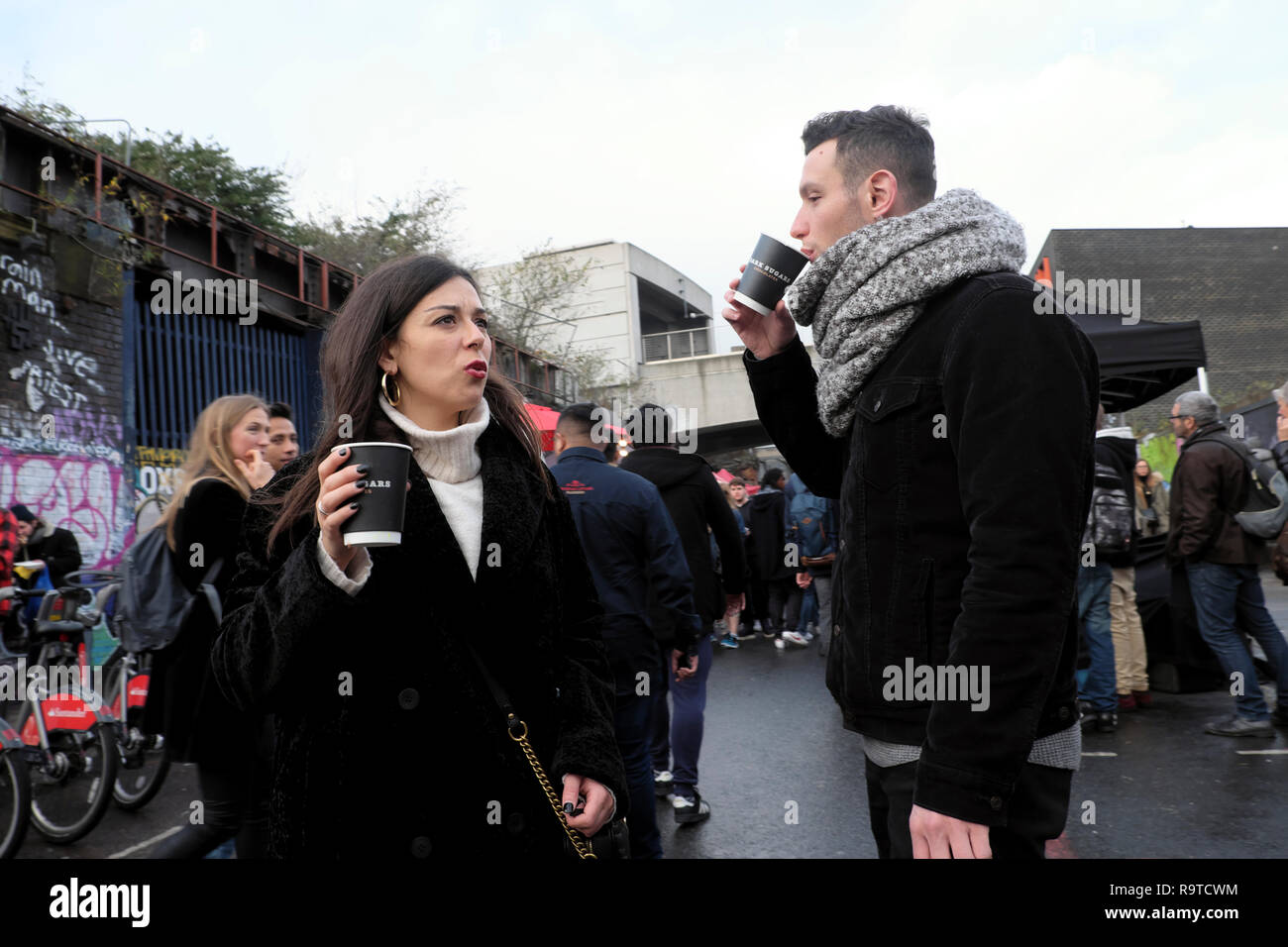People visiting Brick Lane East End young couple standing in the street drinking hot coffee in winter in Shoreditch East London E1 UK  KATHY DEWITT Stock Photo