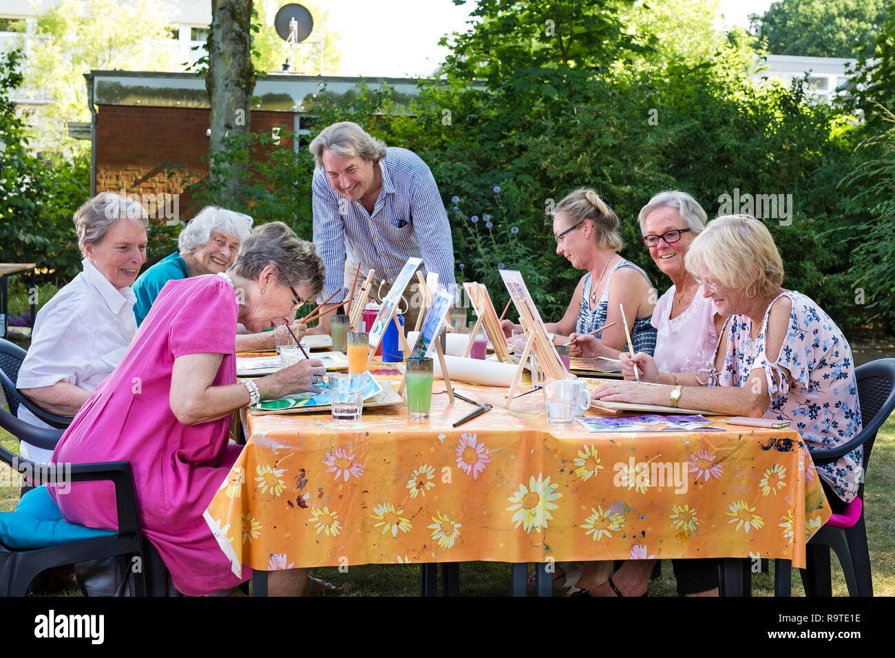 Male artist giving art lessons to the group of senior women, practicing in painting pictures sitting at one table outdoors in backyard. Stock Photo