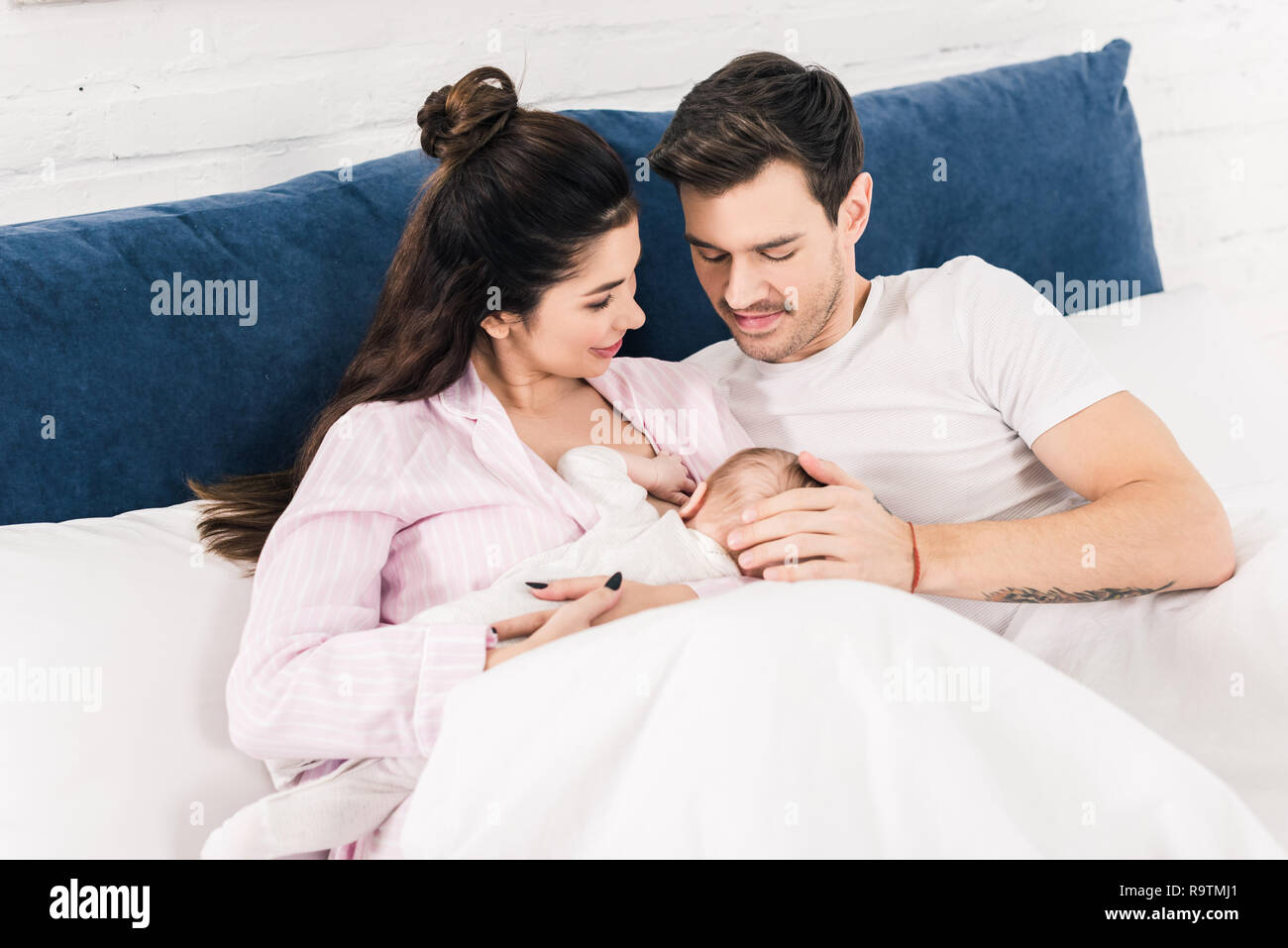 portrait of smiling mother breastfeeding little baby with husband near by on bed at home Stock Photo pic