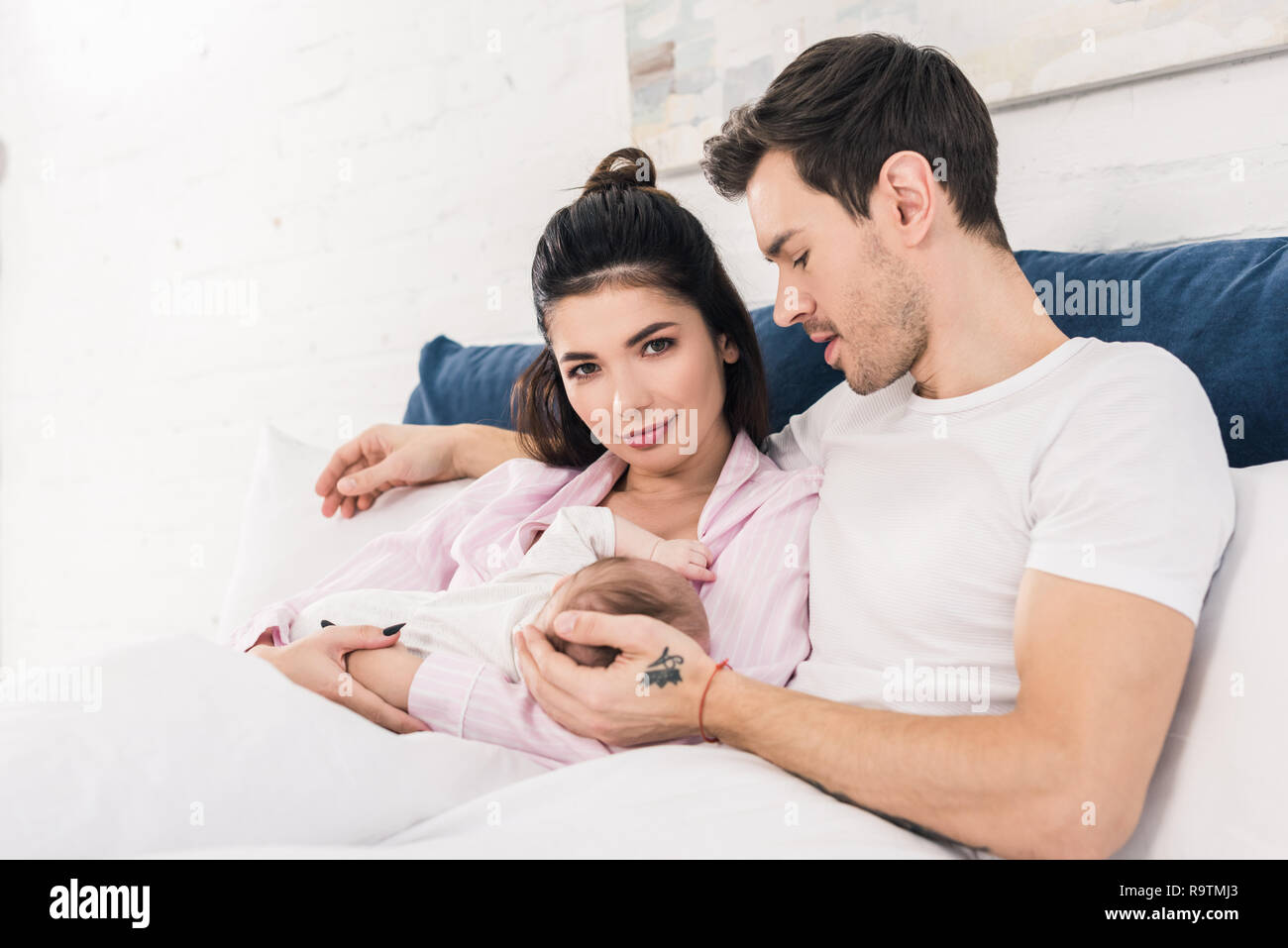 young mother breastfeeding little baby with husband near by on bed at