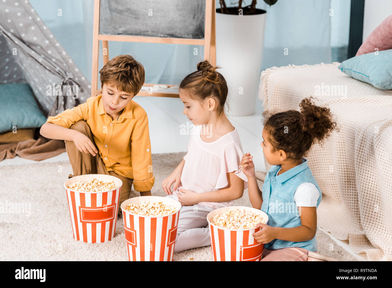 cute little children sitting on carpet and eating popcorn Stock Photo
