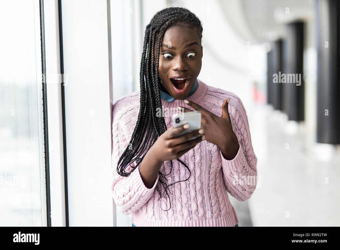 Young surprised afro woman read message from phone in front of panoramic windows Stock Photo