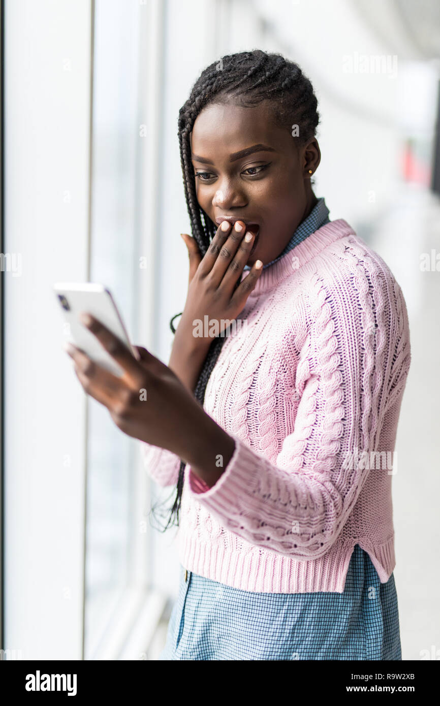 Young surprised afro woman read shocked message from phone in front of panoramic windows Stock Photo