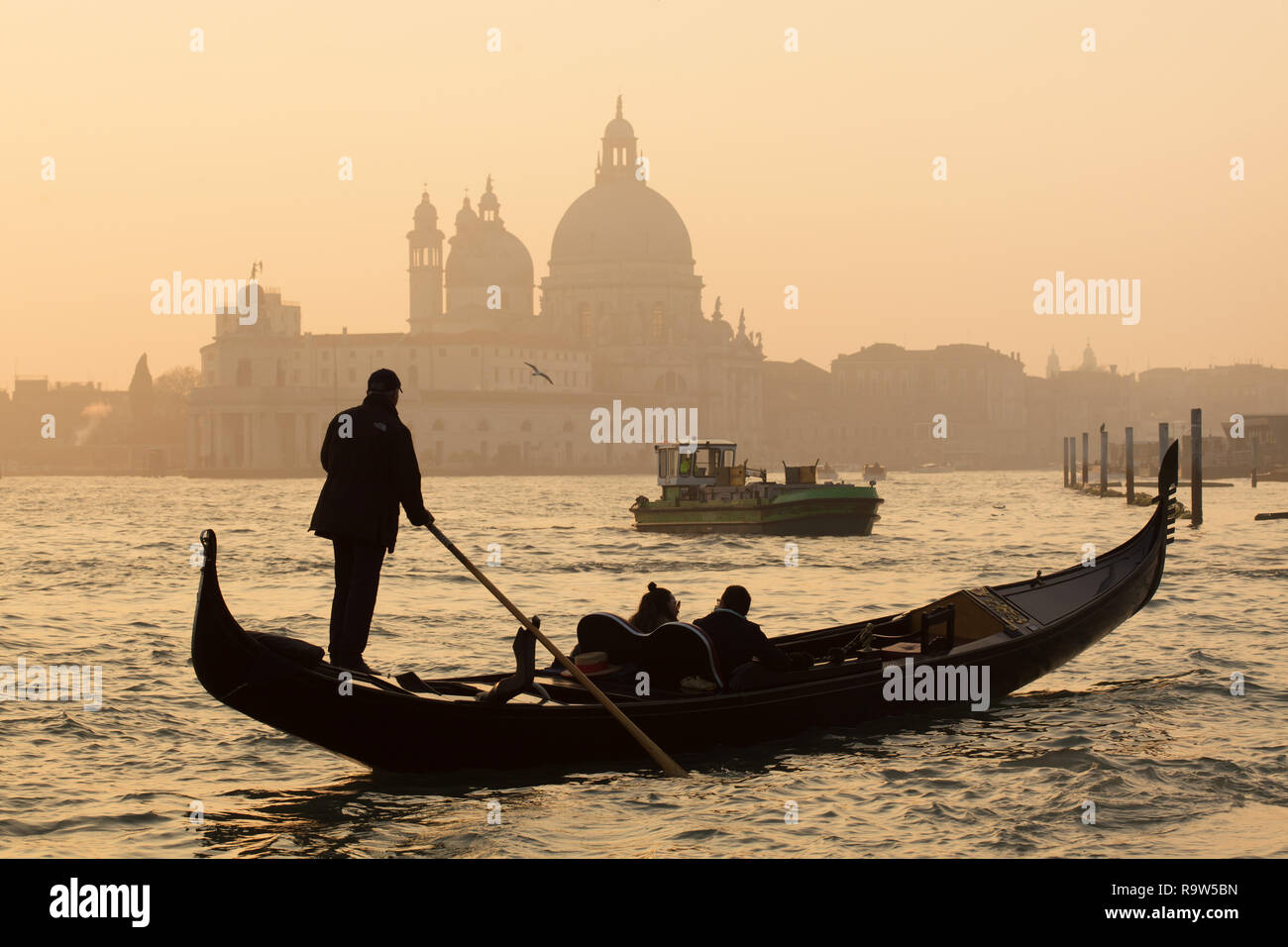 Gondola at sunset in the Venetian Lagoon (Laguna di Venezia) in front of the Basilica of Santa Maria della Salute (Basilica di Santa Maria della Salute) in Venice, Italy. The garbage collecting boat is seen in the background. Stock Photo
