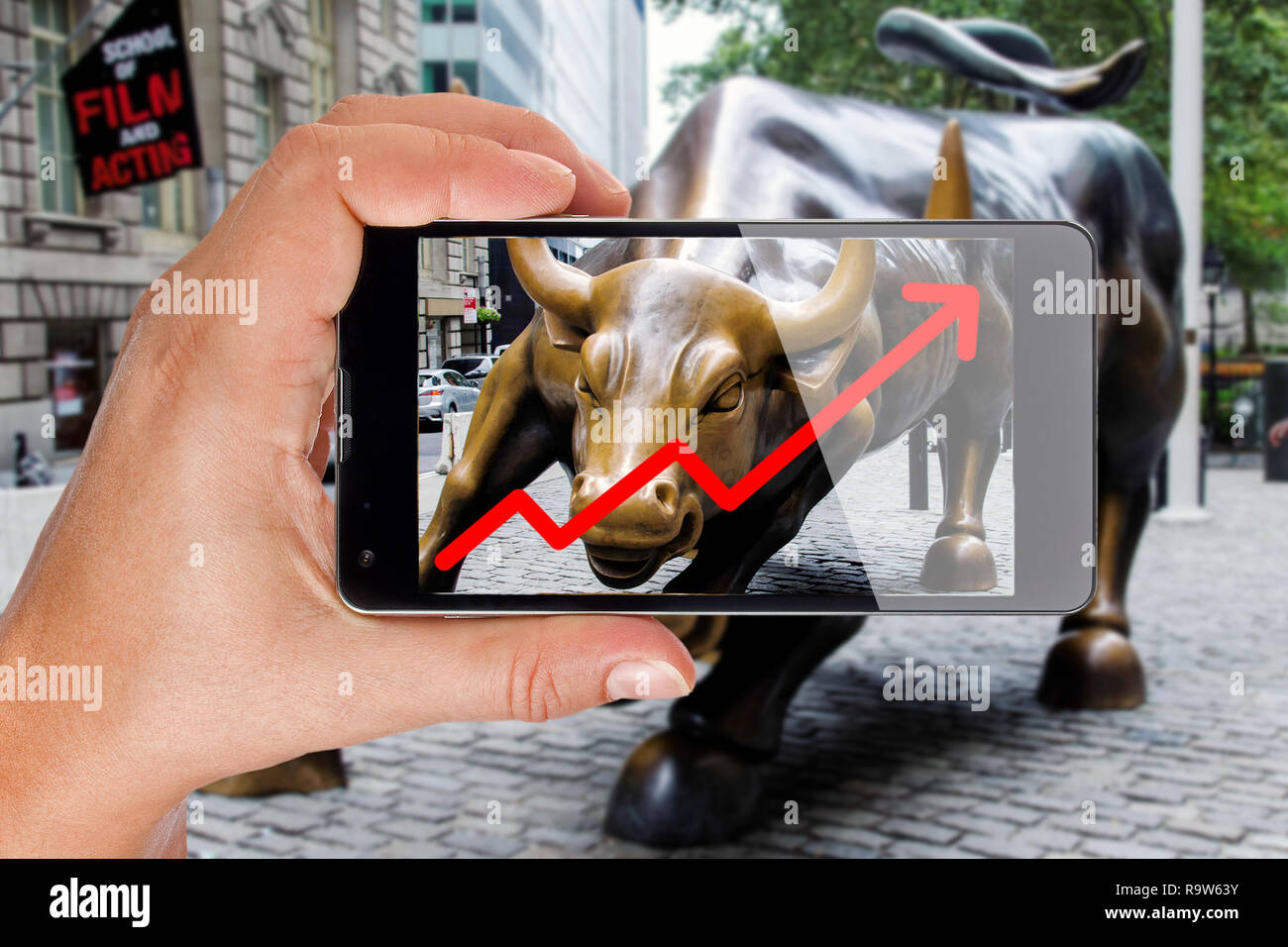 Hand pointing smartphone screen at bull of wall street in New York showing growing stock chart Stock Photo