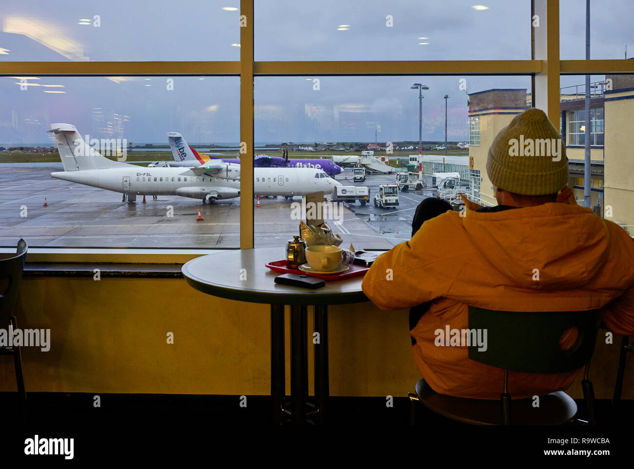 Man wearing woollen hat looking out from Douglas airport cafe at the planes on the tarmac below Stock Photo
