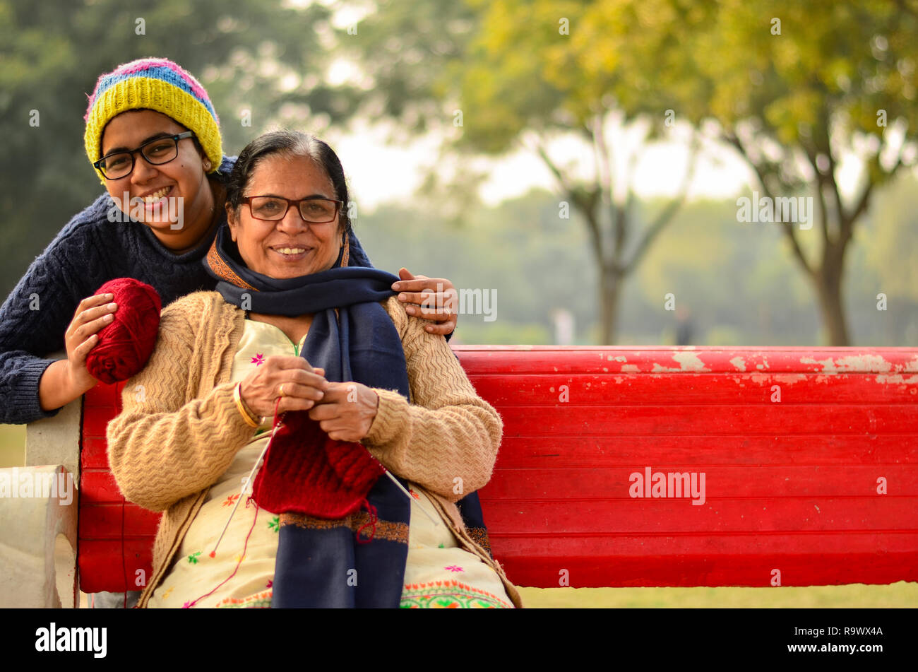 Happy looking young Indian woman with her mother knitting sweater who is sitting on a red bench in a park in Delhi, India Stock Photo