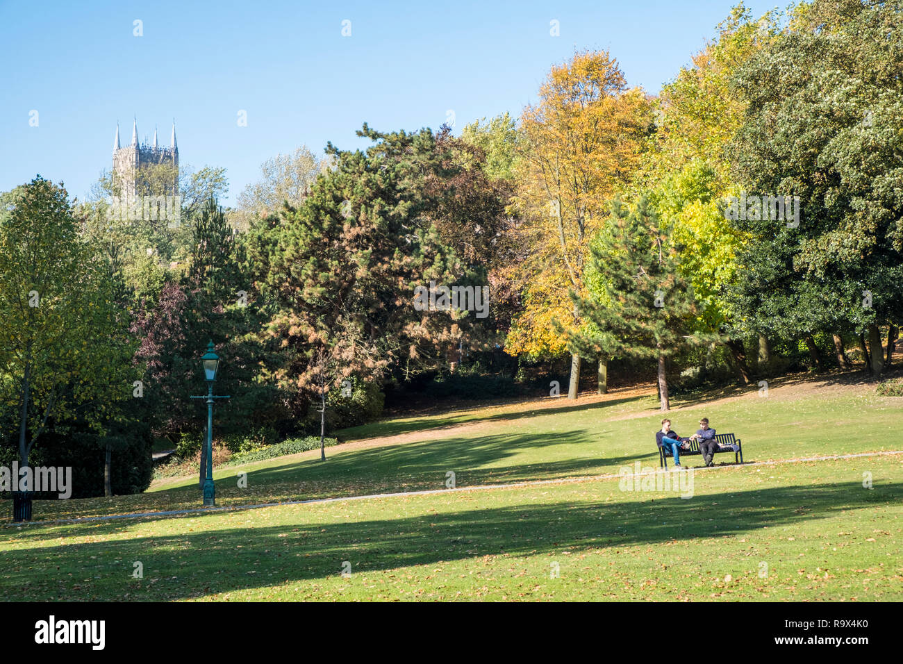 People sitting in a park surrounded by trees in Autumn sunshine. Lincoln Arboretum with Lincoln Cathedral in the distance. Lincoln, England, UK Stock Photo