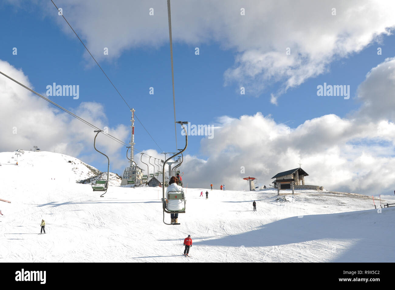 Female in white winter jacket riding a skilift above the slopes, people skiing beneath the lift, a communications tower on top of mountain Stock Photo