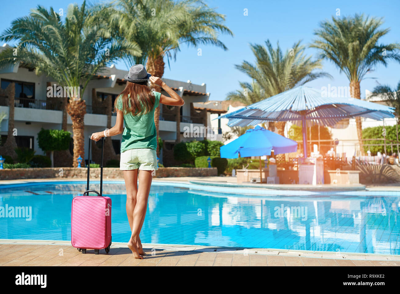 Travel, summer holidays and vacation concept - Beautiful woman walking near exotic hotel pool area with pink suitcase Stock Photo
