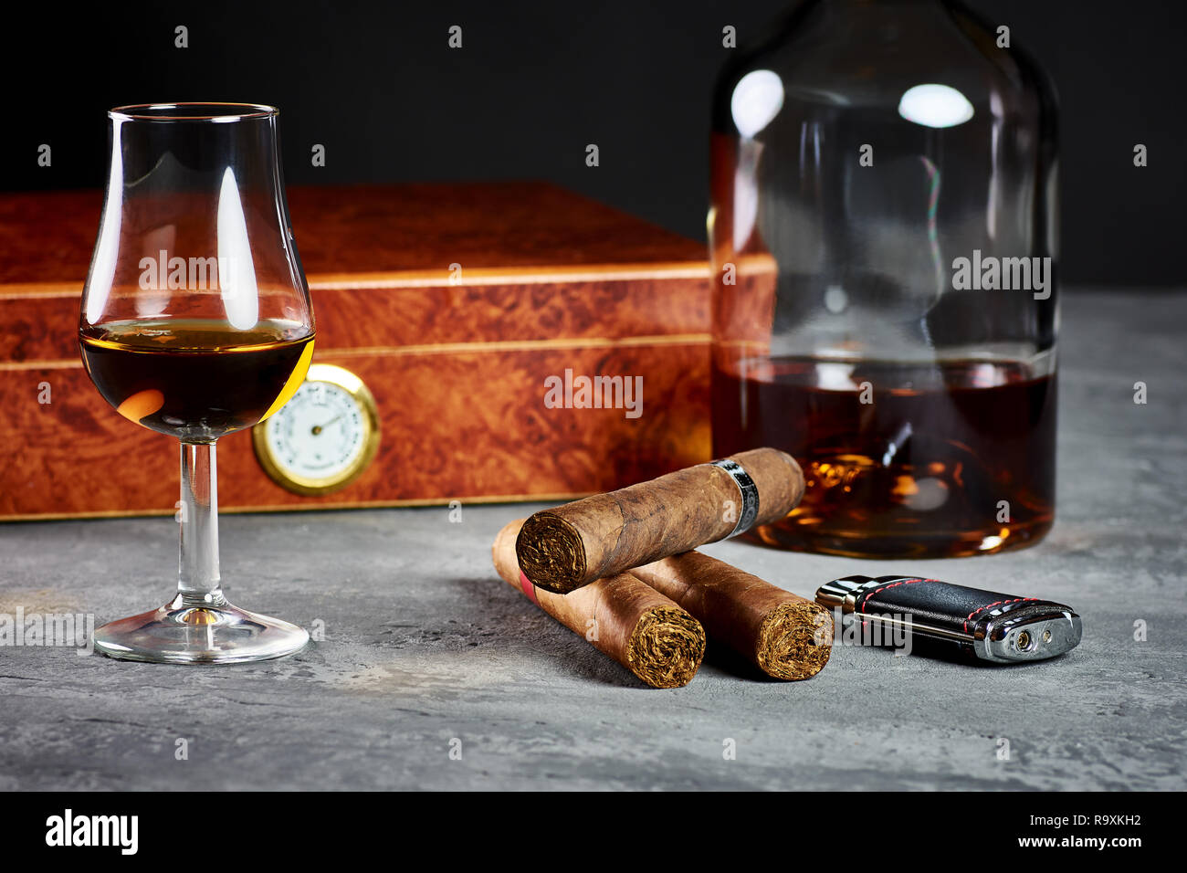 Three Cuban cigars on a stone table with a lighter and a wooden humidor, a glass and a bottle of Whiskey. Stock Photo