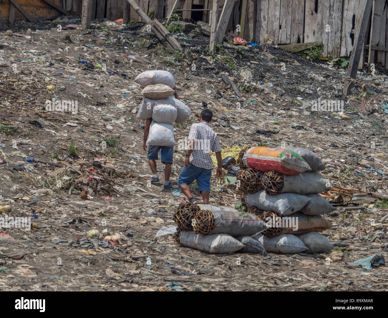 Iquitos, Peru - Sep 25, 2018: Men are carring the bags with charcoal. A huge pollution can be seen in the background on the bank of Itaya River. Low w Stock Photo