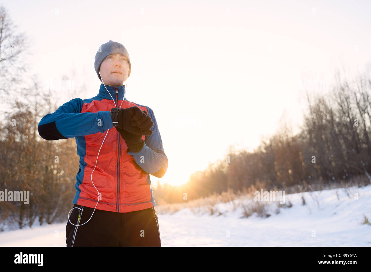 Runner wearing warm sporty clothes in headphones looks at a modern smart clock and counts up spent calories during winter training outdoors Stock Photo