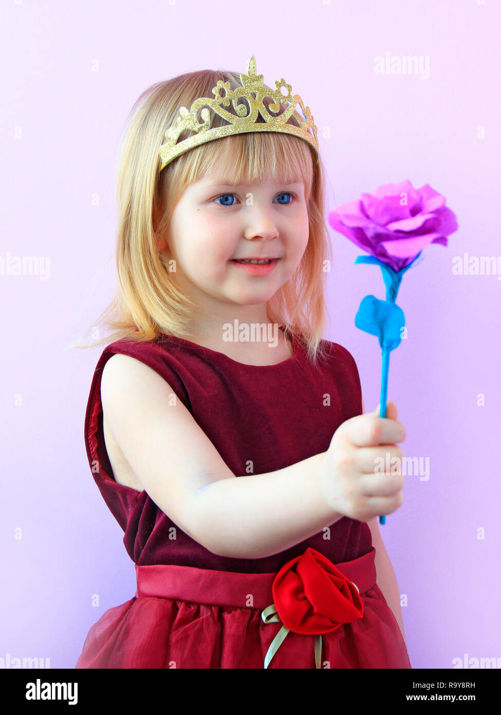 Little girl in crown giving flower. Little princess with pink flower. Cute girl with crown on head offering pink rose. Child in beautiful dress smilin Stock Photo