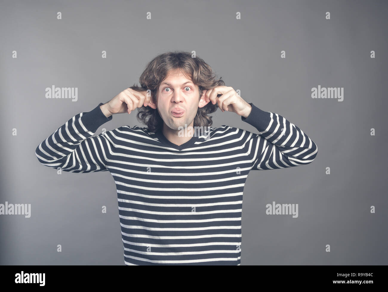 Man making silly monkey face isolated on grey background. Guy grimaces monkey. Funny male holding his ears and shows tongue looks like monkey, pretend Stock Photo