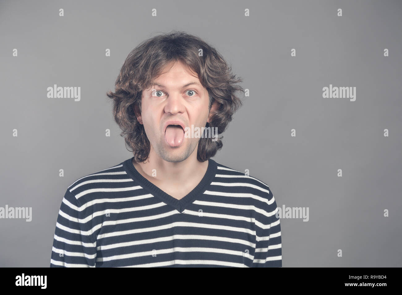 Close up portrait of funny shaggy crazy handsome man in striped sweater looking at camera with big open mouth and tongue out, teases. Wide opened eyes Stock Photo