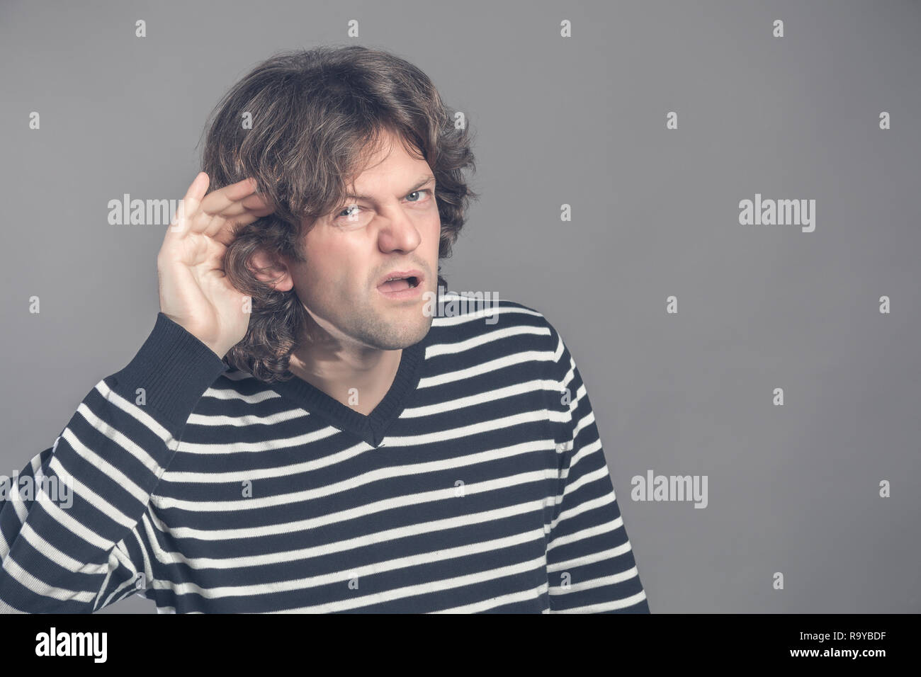 Man with impaired hearing struggling to hear frowning as he holds his hand to his ear in an attempt to improve acoustics. Gossip. Angry man holding ha Stock Photo