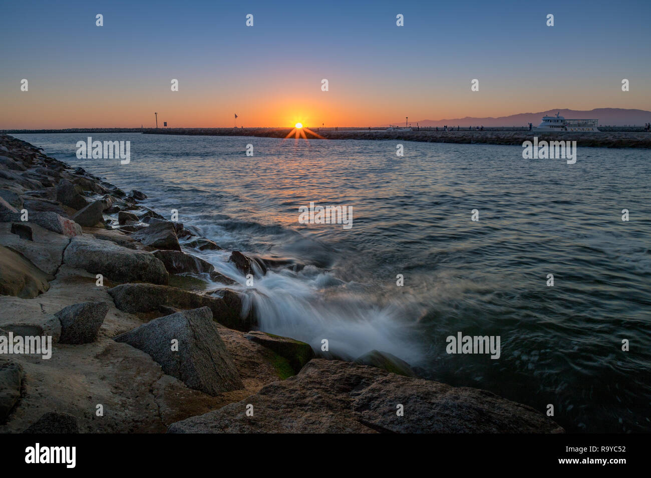 Clear, colorful sky with the sun on the horizon at sunset and waves crashing into a rocky jetty, Marina del Rey, California Stock Photo