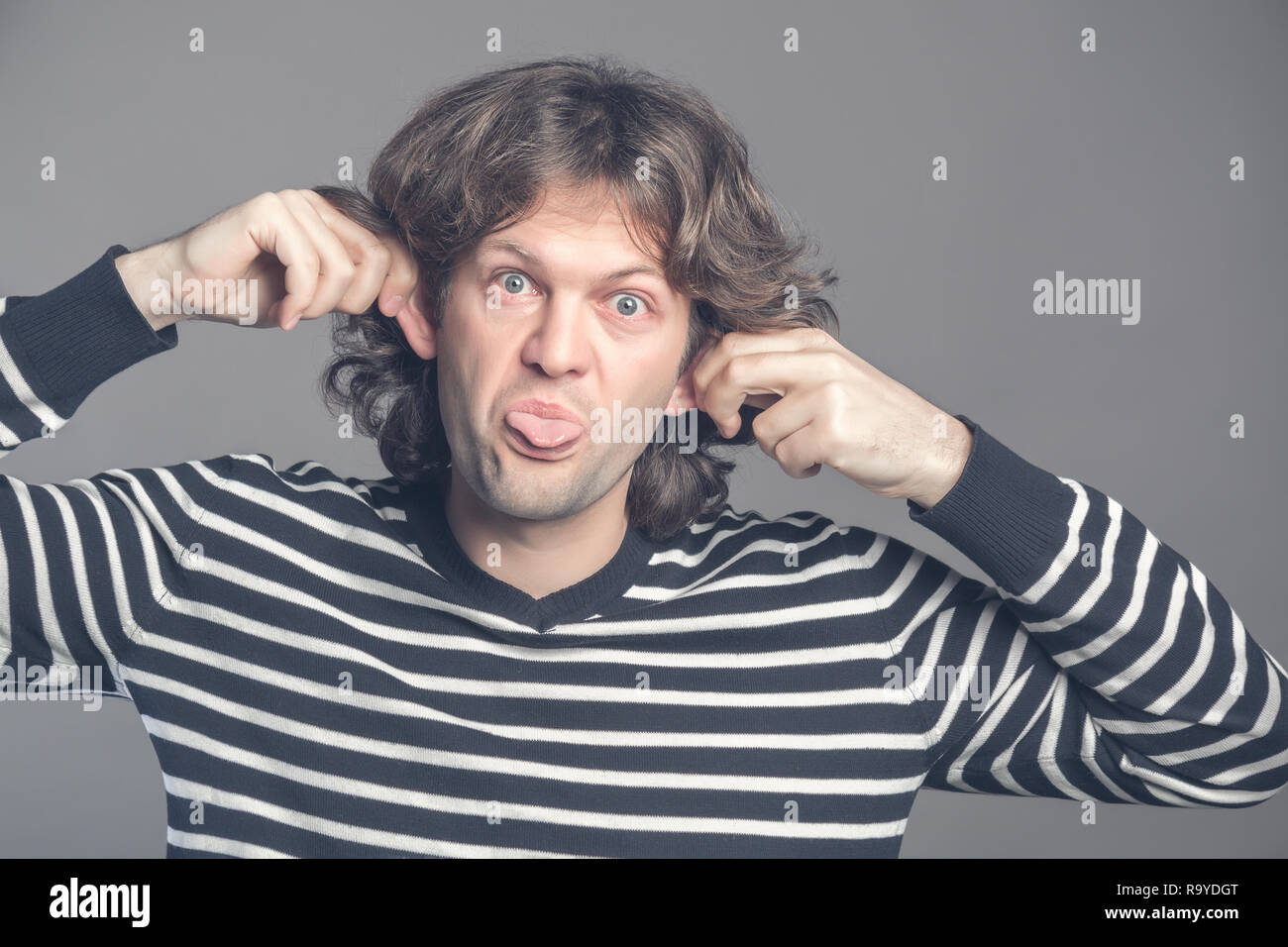Close up of man making silly monkey face isolated on grey background. Guy grimaces monkey. Funny male holding his ears and shows tongue looks like mon Stock Photo