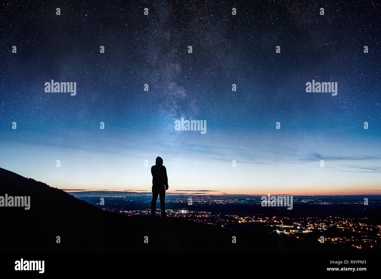 A lone sihoutted hooded figure. Standing on a hill looking down on city lights at night with a galaxy and stars rising in the night sky. Stock Photo