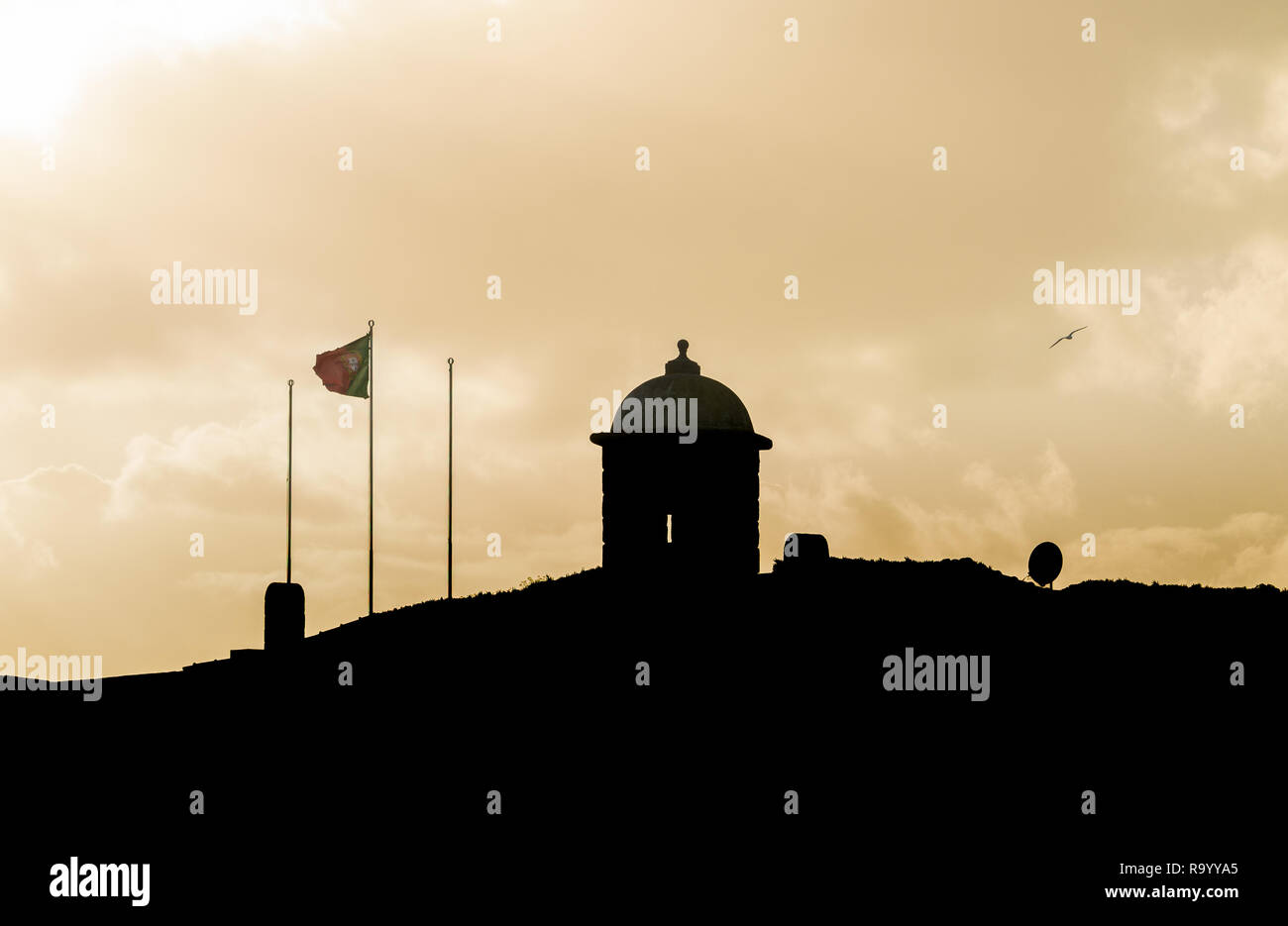 Silhouette of a small castle with a portuguese flag waving in the wind and a seagull flying by. Sunset. Copy space. Stock Photo
