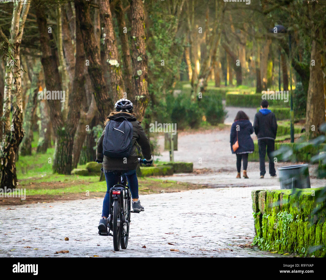Rear view of a young girl riding her bike at a green forest park. Black backpack. Distant people. Stock Photo