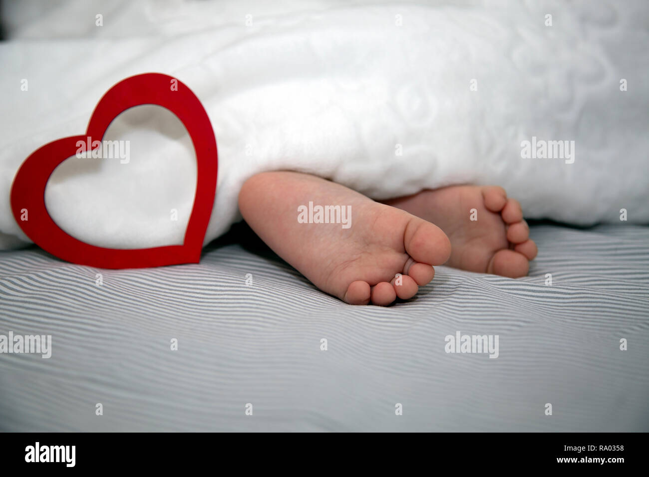 The baby sleeps under a blanket, feet out with a big red heart for the holiday Stock Photo